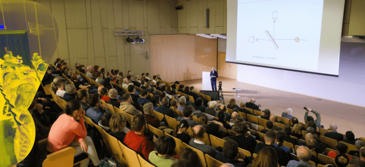  Prelegent prowadzi wykład w dużej auli z ekranem projekcyjnym, sala wypełniona słuchaczami.