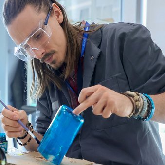  Mężczyzna w okularach ochronnych starannie maluje niebieski szklany słoik.