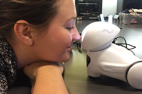 Po lewej stronie widać głowę kobiety, opartą na rękach, nosem styka się z robotem po prawej stronie zdjęcia.