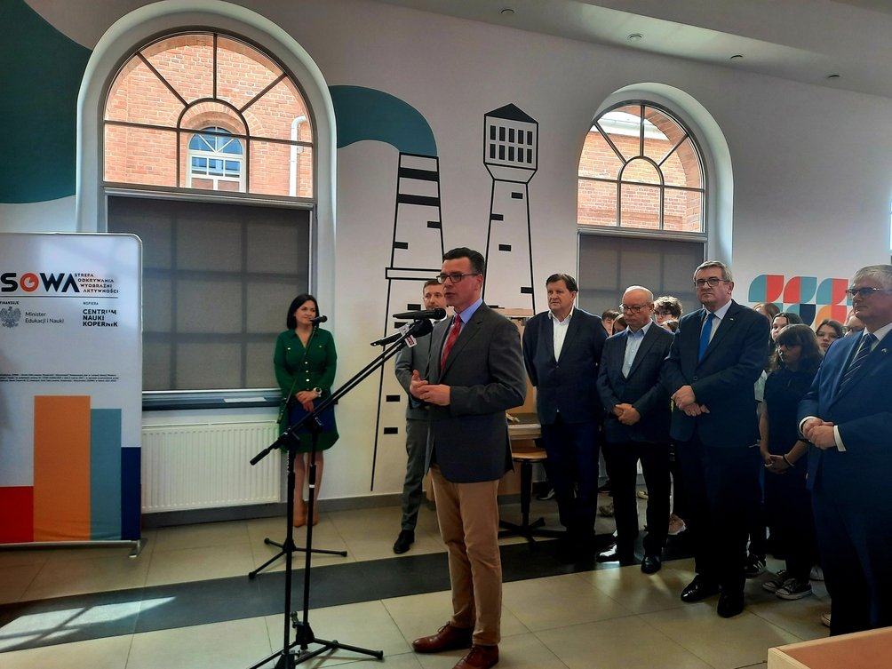 Wnętrze budynku, przy mikrofonie stoi dyrektor Kopernika, za nim wielu ludzi w garniturach, po lewej stronie widać rollup z napisem "SOWA".