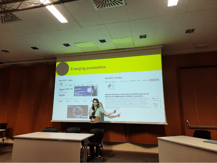 Katarzyna Potęga prezentuje slajd na konferencji w sali z drewnianymi panelami. Slajd zatytułowany „Emerging possibilities” omawia projekty na lata 2023–2024, w tym badania dotyczące społeczeństwa cyfrowego. Kobieta gestykuluje, wskazując na treść.