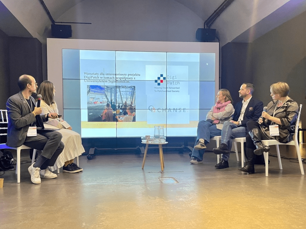 Zdjęcie przedstawia panel dyskusyjny w sali konferencyjnej. Pięciu prelegentów siedzi na scenie na białych krzesłach, z czego dwie osoby po lewej omawiają prezentację wyświetlaną na dużym ekranie w tle. Na ekranie widoczne są zdjęcia z warsztatów oraz logo projektu DigiPatch i programu CHANSE. Atmosfera spotkania jest profesjonalna i skupiona na naukowych dyskusjach.