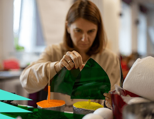 Kobieta skupiona na pracy z kolorowymi foliami i materiałami plastycznymi, tworząc kreatywną konstrukcję na stole. W tle widać rozmyte wnętrze pomieszczenia.