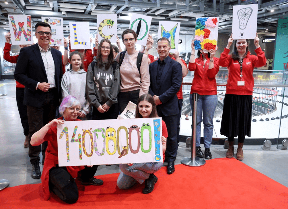 Zdjęcie przedstawia grupę ludzi pozujących na czerwonym dywanie w nowoczesnym budynku. W tle widać przestrzeń wystawową oraz instalacje techniczne. Osoby w czerwonych bluzach trzymają ozdobne tabliczki układające się w napis „WELCOME”. Na pierwszym planie dwie kobiety trzymają duży, kolorowy napis „14 000 000”, ozdobiony rysunkami zwierząt i wzorami. W centrum grupy znajdują się dwie dziewczynki oraz dwaj mężczyźni w eleganckich strojach.