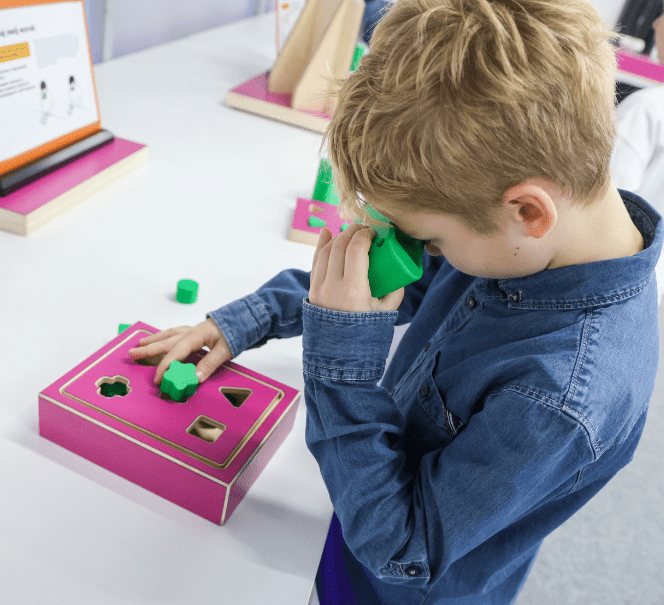 Chłopiec przykłada zielony klocek do oka i dopasowuje kształty do otworów w różowym stanowisku, ćwicząc spostrzegawczość i wyobraźnię przestrzenną.