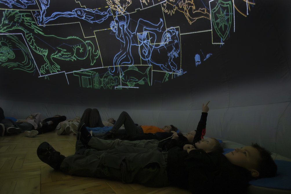 Wnętrzne mobilnego planetarium, dość ciemno, na podłodze leżą dzieci i pokazują ręką coś na kopule.