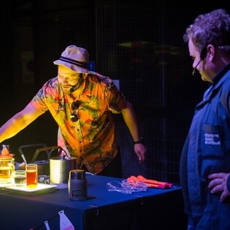 Dwoje ludzi w ciemnym pomieszczeniu, jeden z mężczyzn jest pochylony nad stołem i przelewa coś do kolorowych zlewek.