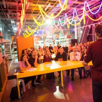 Kolorowe pomieszczenie ze stołami, pod sufitem wiszą kolorowe ledy, widać publiczność, która słucha prowadzącego.