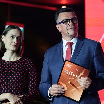  Prowadzący galę Nagrody Przewroty trzymają teczki z logo wydarzenia, stojąc na scenie.