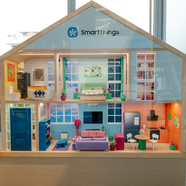 Makieta inteligentnego domu SmartThings od Samsunga, przedstawiona w formie przekroju przez ściany. Domek ma dwa piętra z widocznymi pomieszczeniami: łazienką, sypialnią, garderobą, salonem, kuchnią i przedpokojem. W każdym pomieszczeniu znajdują się nowoczesne, miniaturowe meble i kolorowe dekoracje. Na dachu widnieje logo SmartThings. Całość przypomina realistyczny dom.