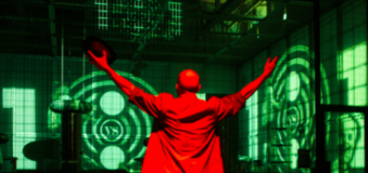 Ciemne pomieszczenie, na środku stoi tyłem podświetlona na czerwono osoba z uniesionymi rękami.