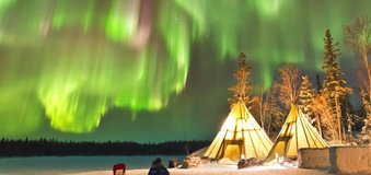  Zorza polarna tańcząca nad zimowym obozowiskiem z tipi i osobą siedzącą na śniegu.