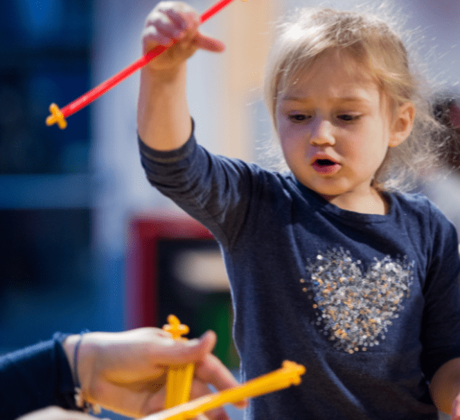 Dziewczynka w granatowej bluzki, w uniesionej rece trzyma zabawkę.