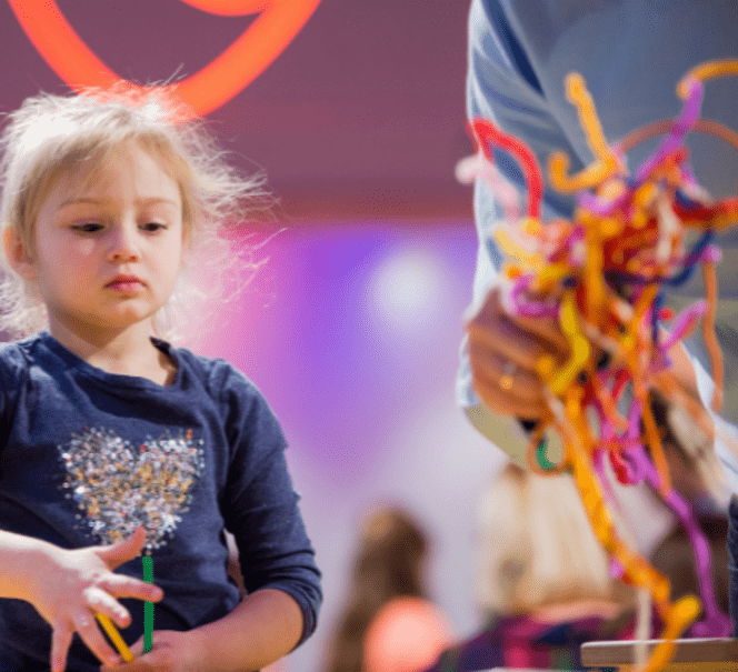 W lewej części zdjęcia widać dziewczynkę, z prawej strony, bliżej widać dorosłego człowieka trzymającego naręcze kolorowych drucików.