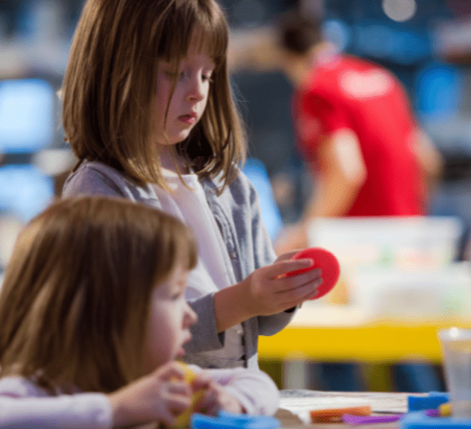 Dwie dziewczynki, jedna z nich trzyma czerwone kółko, w tle widać stoły z pudełkami oraz animatora.