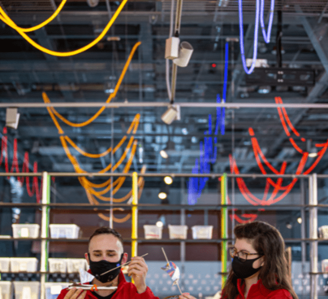 Większość kadru zajmują kolorowe światła pod sufitem, niżej widać dwoje animatorów trzymających samodzielnie zbudowane łódki. z patyczków.