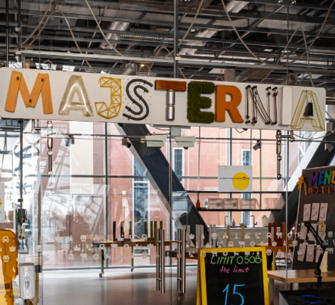 Zdjęcie wejścia do Majsterni, napis jest złożony z różnych elementów jak deski, światła led, mech...