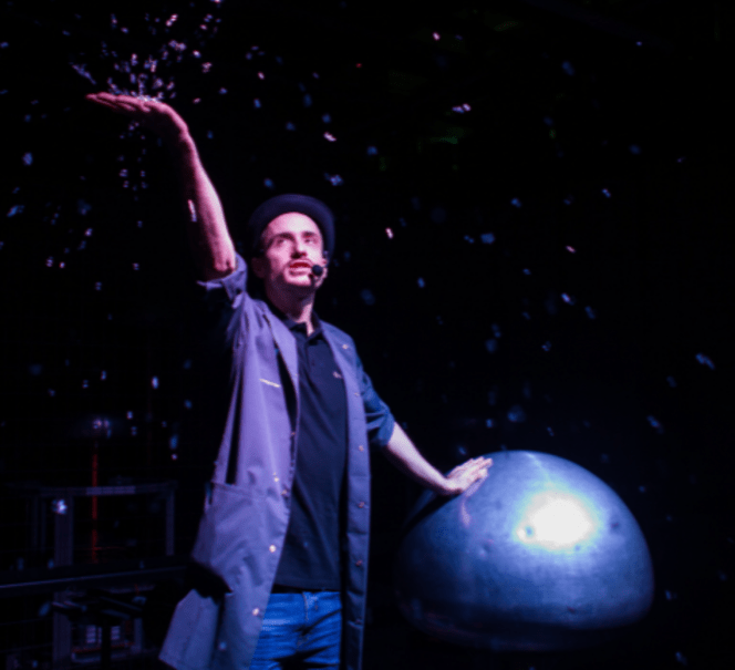 Animator w szarym fartuchu, jedną rękę trzyma na wielkiej metalowej kuli, drugą wyrzuca w powietrze kolorowe papierki.