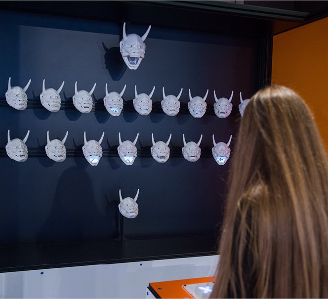 Widać tył głowy kobiety, która stoi przed ścianą pełną twarzy demonów.