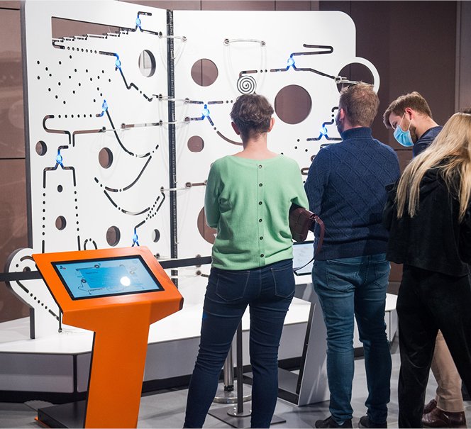 Ludzie tyłem, stoją i wpatrują się w eksponat - ścianę z wyciętymi dziurami.