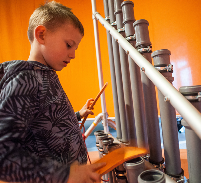 Chłopiec uderzający czymś w instalację z rurek pcv, która przypomina instrument muzyczny.