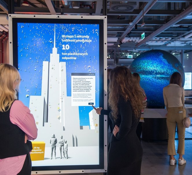 Na pierwszym planie widać dwie kobiety wpatrzone w ekran z informacją o ilości produkowanych śmieci na świecie, na drugim planie jest eksponat przypominający kulę ziemską.