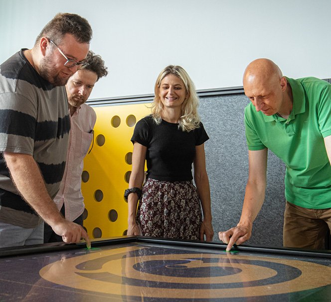 Troje ludzi pochyla się nad stołem, na którym są widoczne koliste malunki.