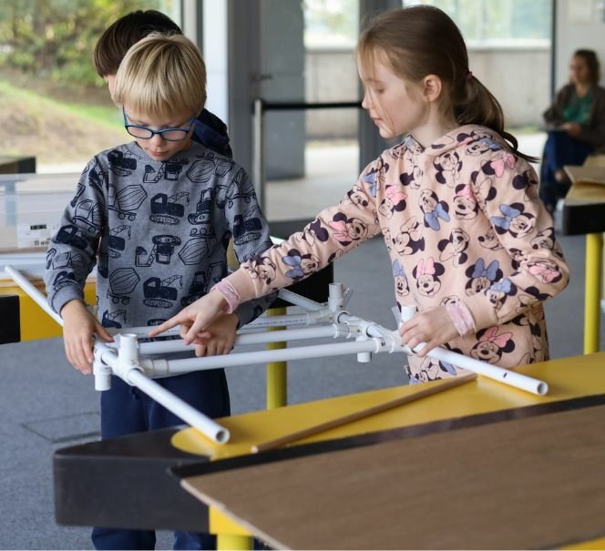 Dwoje dzieci w bluzach z nadrukami buduje konstrukcję z plastikowych rurek przy stole w przestrzeni edukacyjnej. Chłopiec w okularach trzyma elementy, a dziewczynka wskazuje palcem na połączenie. W tle inni uczestnicy zajęć.