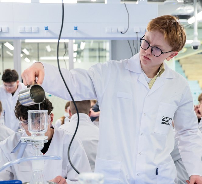  Uczeń w białym fartuchu i okularach ochronnych wlewa ciecz do zestawu laboratoryjnego.