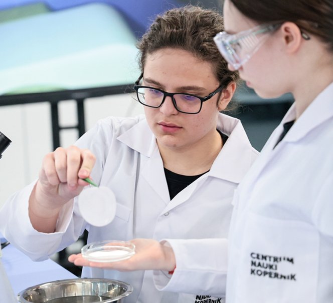  Dwoje uczniów w fartuchach pracuje z naczyniami laboratoryjnymi, jedna trzyma pincetą krążek filtra.