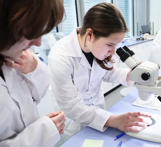  Dziewczyna w białym fartuchu obserwuje próbkę pod mikroskopem, obok niej druga uczennica, obie skupione na eksperymencie.