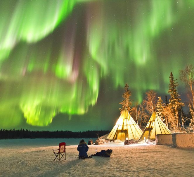  Zorza polarna tańcząca nad zimowym obozowiskiem z tipi i osobą siedzącą na śniegu.