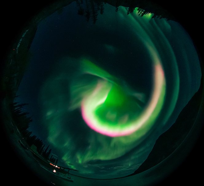  Zorza polarna w formie spiralnego wiru widziana przez obiektyw typu „rybie oko”; dominują zielenie i róże.