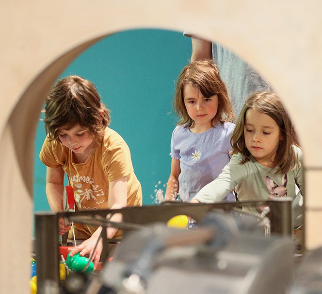 Grupa dzieci pochyla się nad długą instalacją wodną z kolorowymi piłeczkami.