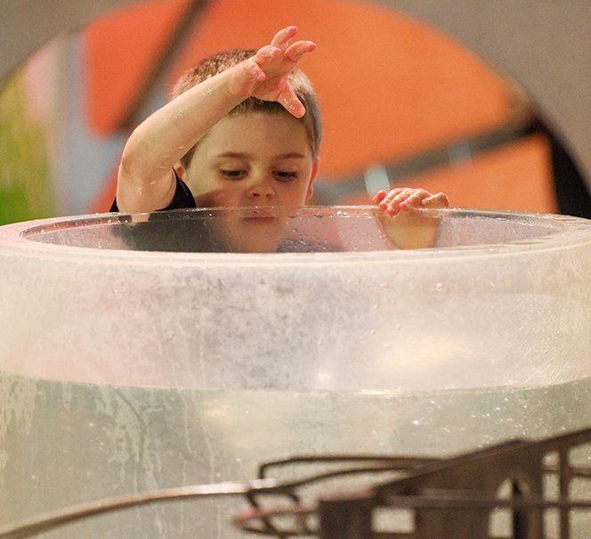 Chłopiec wrzuca coś do przezroczystego zbiornika z wodą, w tle wystawa.