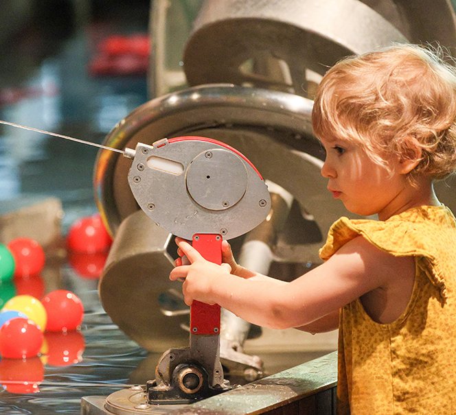 Dziewczynka obraca korbkę w instalacji z kolorowymi piłkami w wodzie.