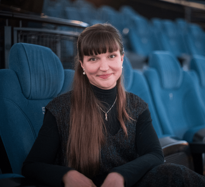 Kobieta z długimi, ciemnymi włosami i grzywką uśmiecha się, siedząc na niebieskim fotelu w planetarium.