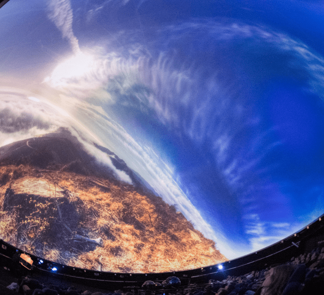 W planetarium wyświetlana jest projekcja krajobrazu z niebem i chmurami; widzowie patrzą w górę.
