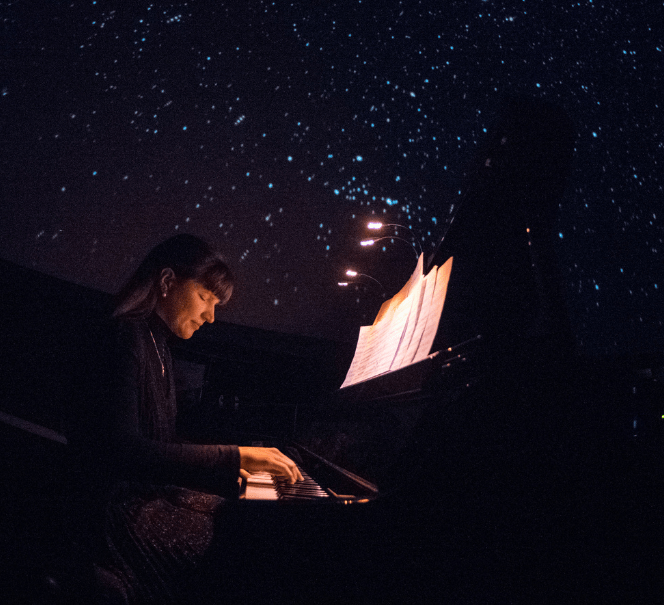Pianistka gra na fortepianie w ciemnym planetarium, nad nią rozgwieżdżone niebo wyświetlone na kopule.