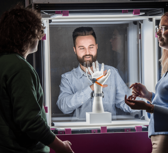Trzy osoby obserwują sztuczną dłoń w szklanej gablocie.
