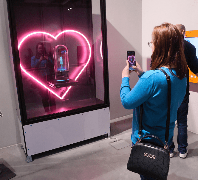 Osoba fotografuje neonowe serce z hologramem postaci w środku.