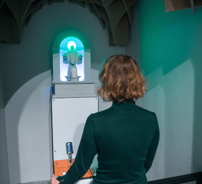 Kobieta tyłem do nas stoi i patrzy na świecącą figurę w kaplicznej scenerii.