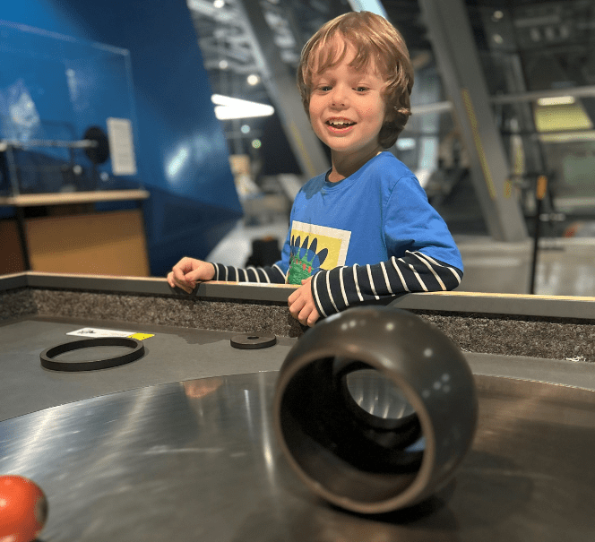 Chłopiec uśmiecha się przy interaktywnym stanowisku z kulkami i rurami w centrum nauki.