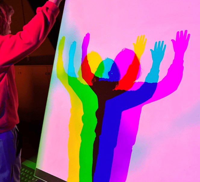 Osoba stoi przed kolorowym ekranem, na którym cień jej sylwetki rozszczepia się na barwy tęczy.