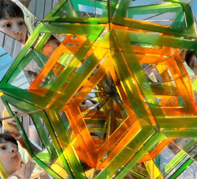 Dziecko zagląda w kolorową konstrukcję z luster i przezroczystych kształtów geometrycznych.